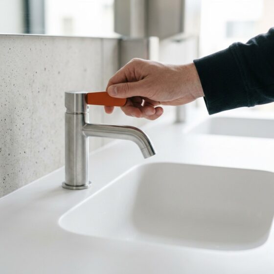 Hand draait aan een kraan met oranje hendel boven een droog wit bassin waaruit geen water komt