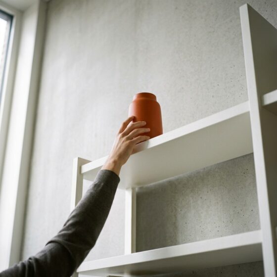 Hand reikt naar een oranje vaas op de hoogste plank van een witte kast, net buiten bereik