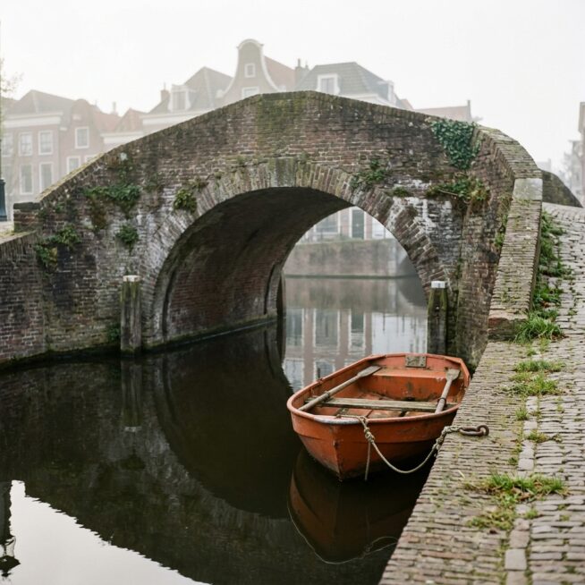 Oude bakstenen boogbrug over een Nederlandse gracht met een oranje roeiboot in de ochtendmist, als beeld voor linkbuilding die verbindt